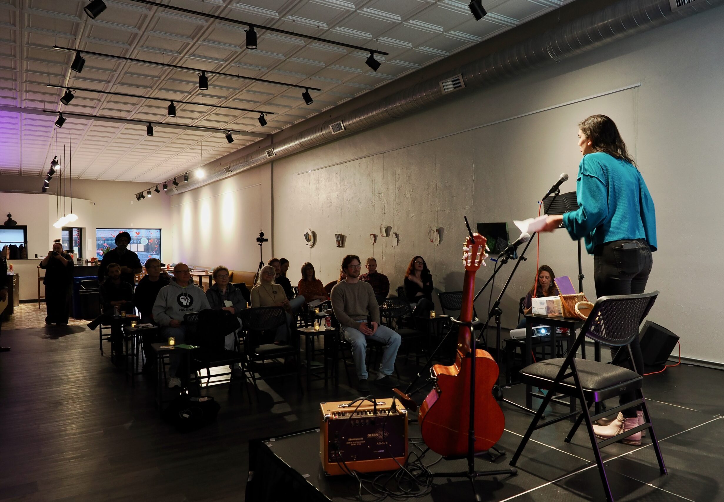 A person in a blue sweater speaks on stage in front of a group of 18 people in a storefont.