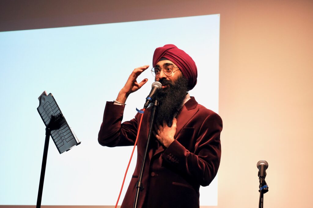 A man in a red turban and vibrant red velvet jacket speaks on stage, with his hand on his heart.