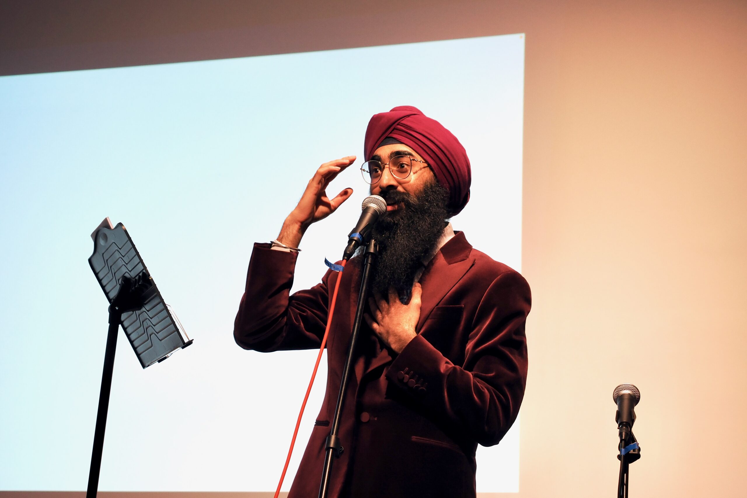 A man in a red turban and vibrant red velvet jacket speaks on stage, with his hand on his heart.