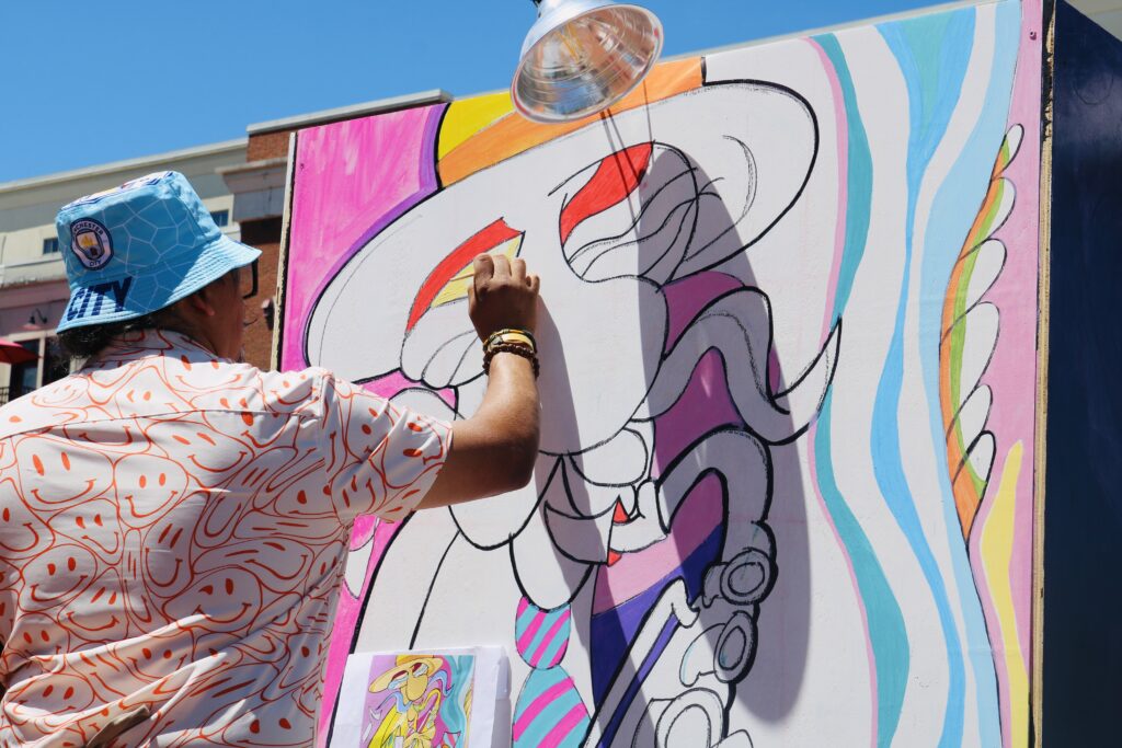 An artist wearing a blue bucket hat and patterned shirt paints bright colors onto a large abstract mural outdoors under a sunny blue sky.