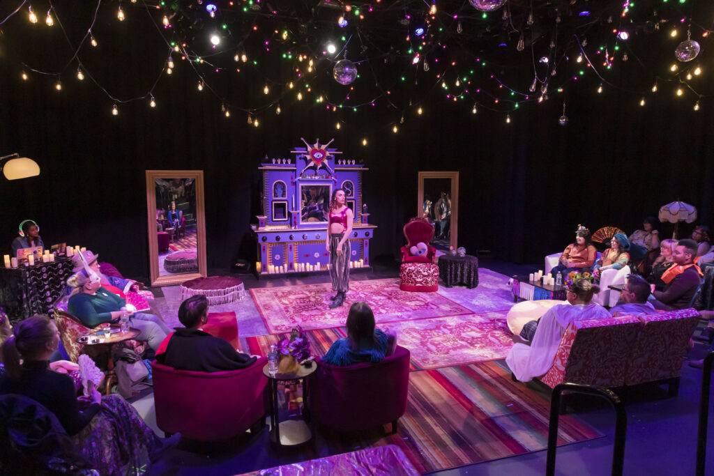 An actor stands in the middle of the floor in a dark theater space, with rugs underfoot and multicolored string lights overhead. Audience members sit on sofas and armchairs, some wearing ornate masks.