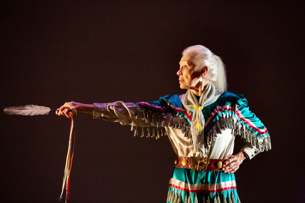 Female dancer looking to the left and pointing with a feather against a black background.