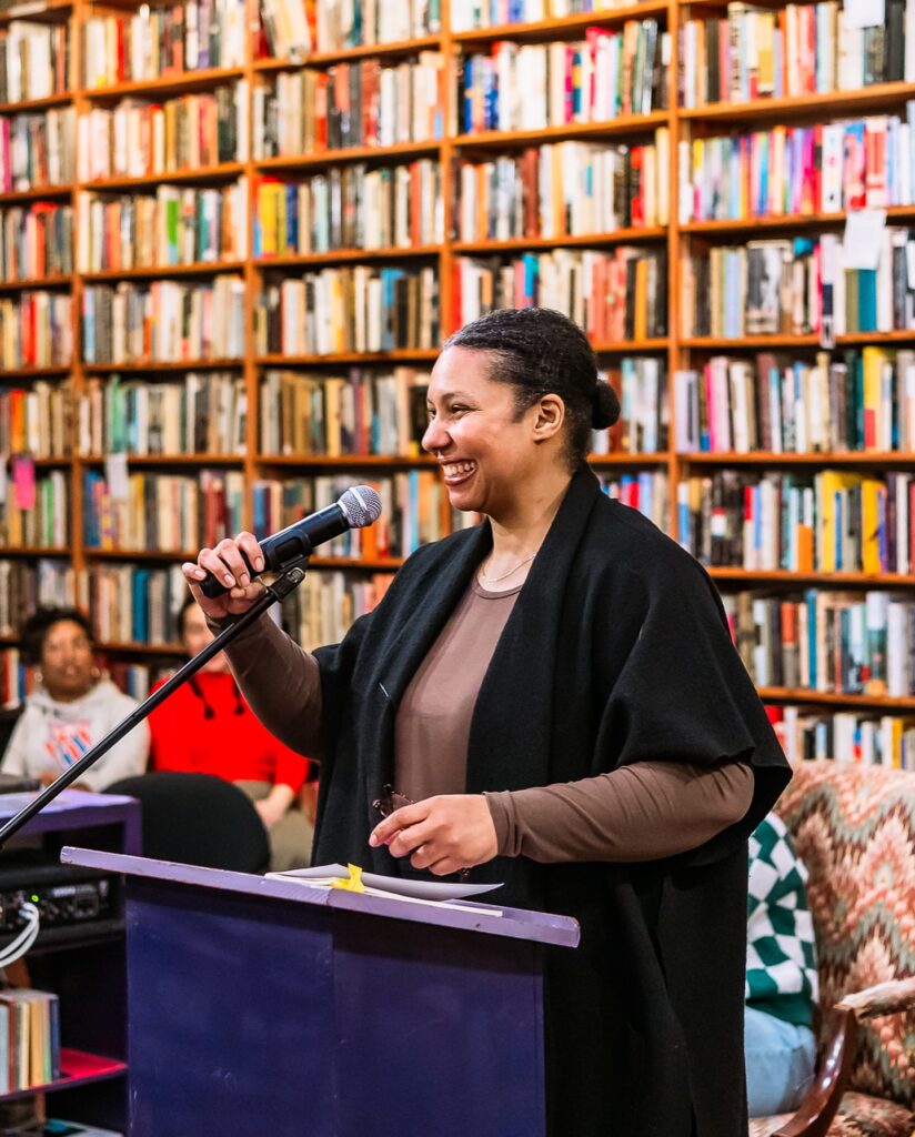 A woman standing in front of a large shelf of books holds a microphone attached to a lectern and smiles at an audience.