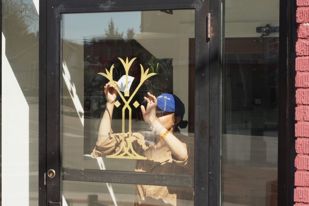A person with medium-dark skin tone wears a blue baseball cap and holds hands up to a gold design on a window.