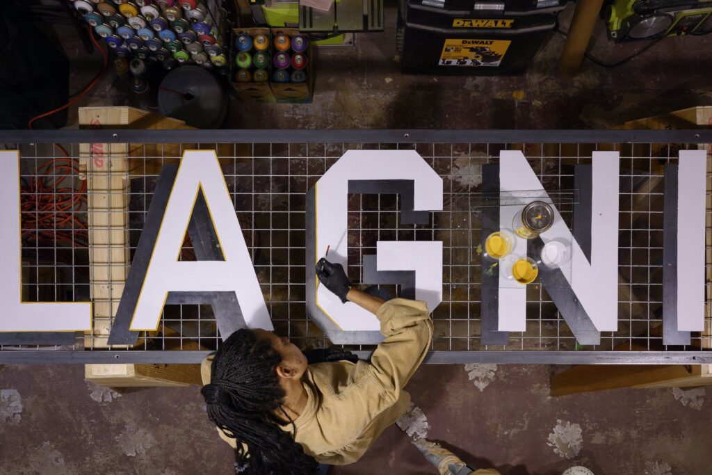 An overhead image of a person with medium-dark skin tone wearing a beige shirt paints a large white letter "G."