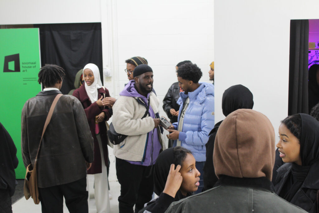 Attendees gather at the center of the gallery space following an artist talk at Soomaal House in Minneapolis, MN. The white walls, in frame, surrounding them have small to medium-sized two-dimensional works of art.