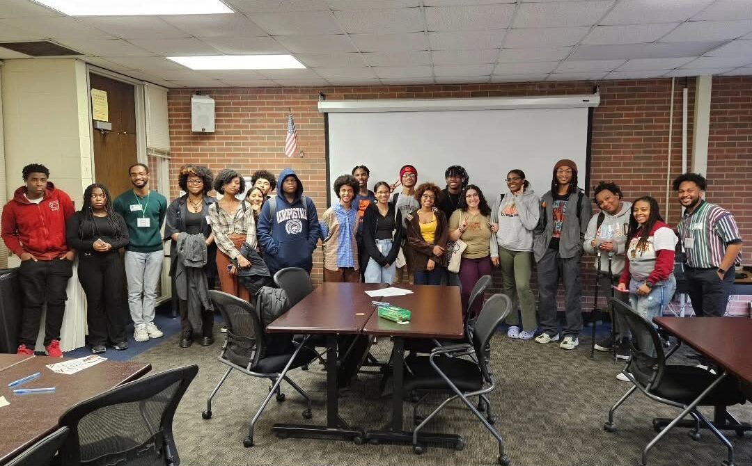 A large group of young adults stand together in a classroom, posing for a group photo in front of a blank projection screen and an American flag. Several tables and chairs are in the foreground.