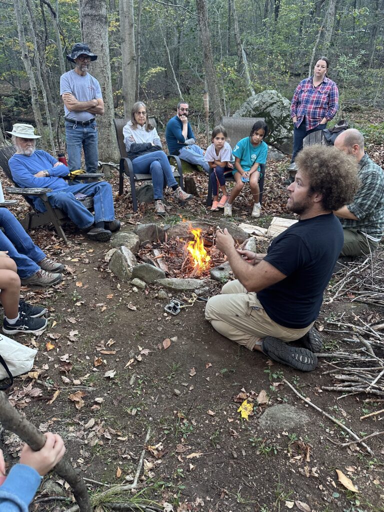 People of different ages sitting outdoors on chairs. They are all looking at a person kneeling on the ground talking to them as they sit by a small fire.