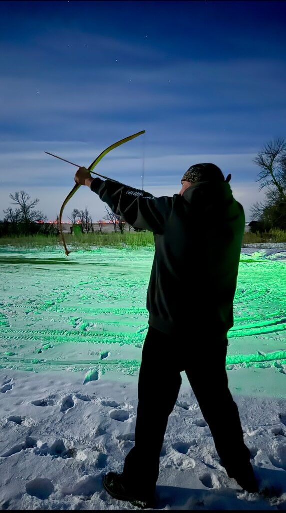 A person standing outside in a snowy yard at night holding a bow and arrow.