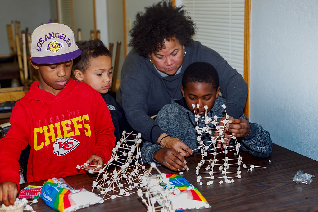 A person of medium skin tone with short curly hair helping several young students make structures with toothpicks and marshmallows.