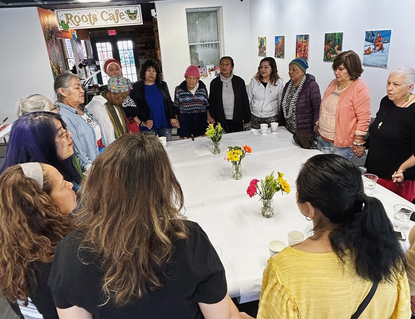 A group of women with various skin tones stand in a circle around a table holding hands. Many appear to be singing.