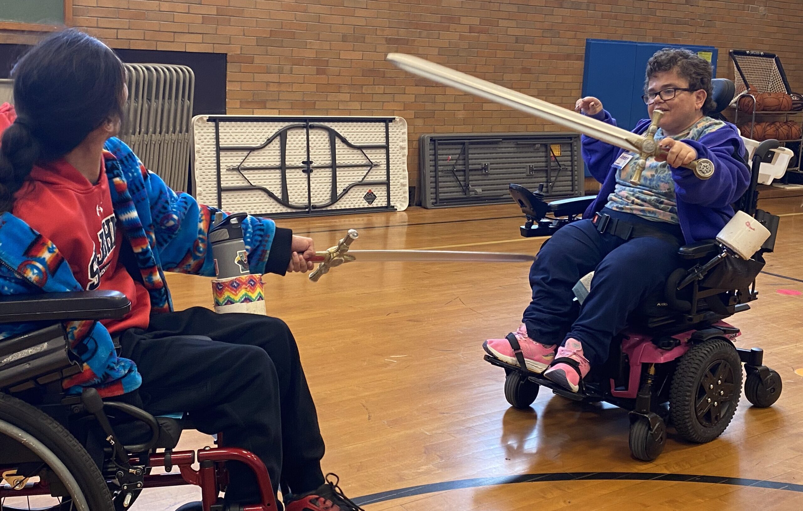 Two people sitting in wheelchairs sword fight in a gymnasium.