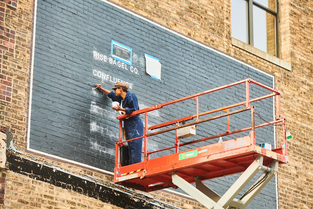 A person with medium-dark skin tone wears a jumpsuit and stands on an orange scissor lift and paints white lettering on a brick building.