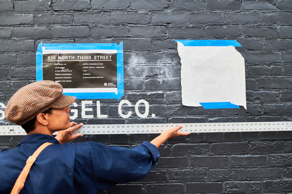 A person with medium-dark skin tone holds a large ruler up against a black brick wall under white lettering and two pieces of taped paper.