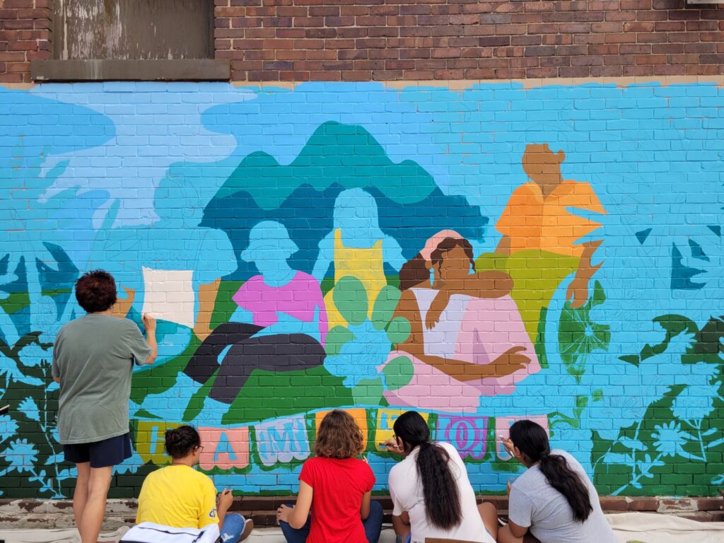 An adult and four youth painting in a mural of five people in front of a mountain.