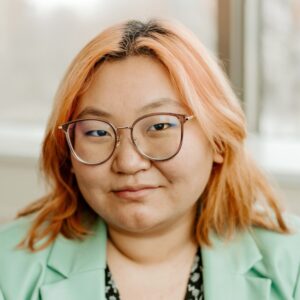 A smiling person of medium light skin tone, with light brown rimmed glasses and shoulder length pink hair, wearing a seafoam green blazer over a black and seafoam green patterned top