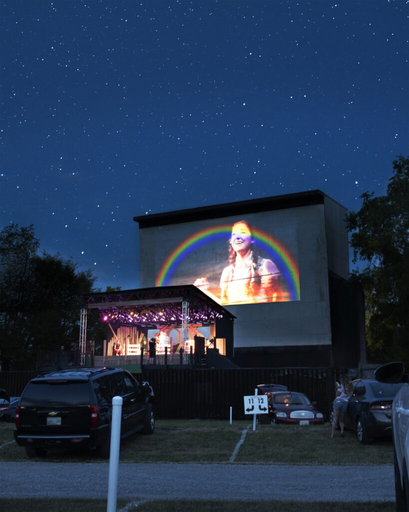A theatre performance on a small stage being projected onto a big screen at a drive-in movie theater.