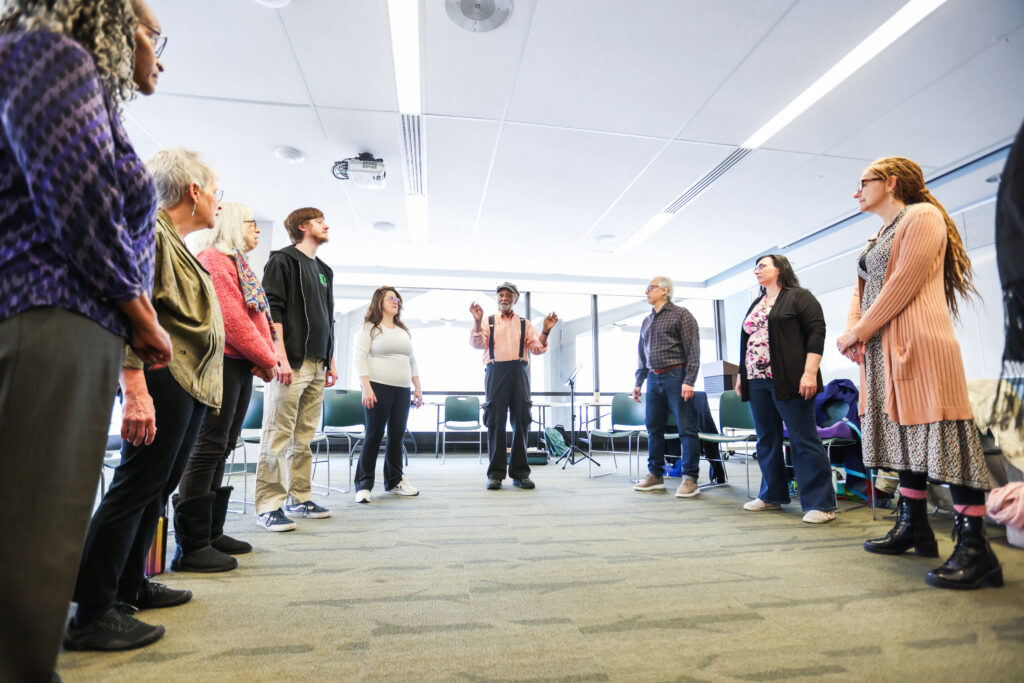 A group of adults stand in a circle in a bright meeting room, listening to a facilitator in the center who gestures as he speaks.