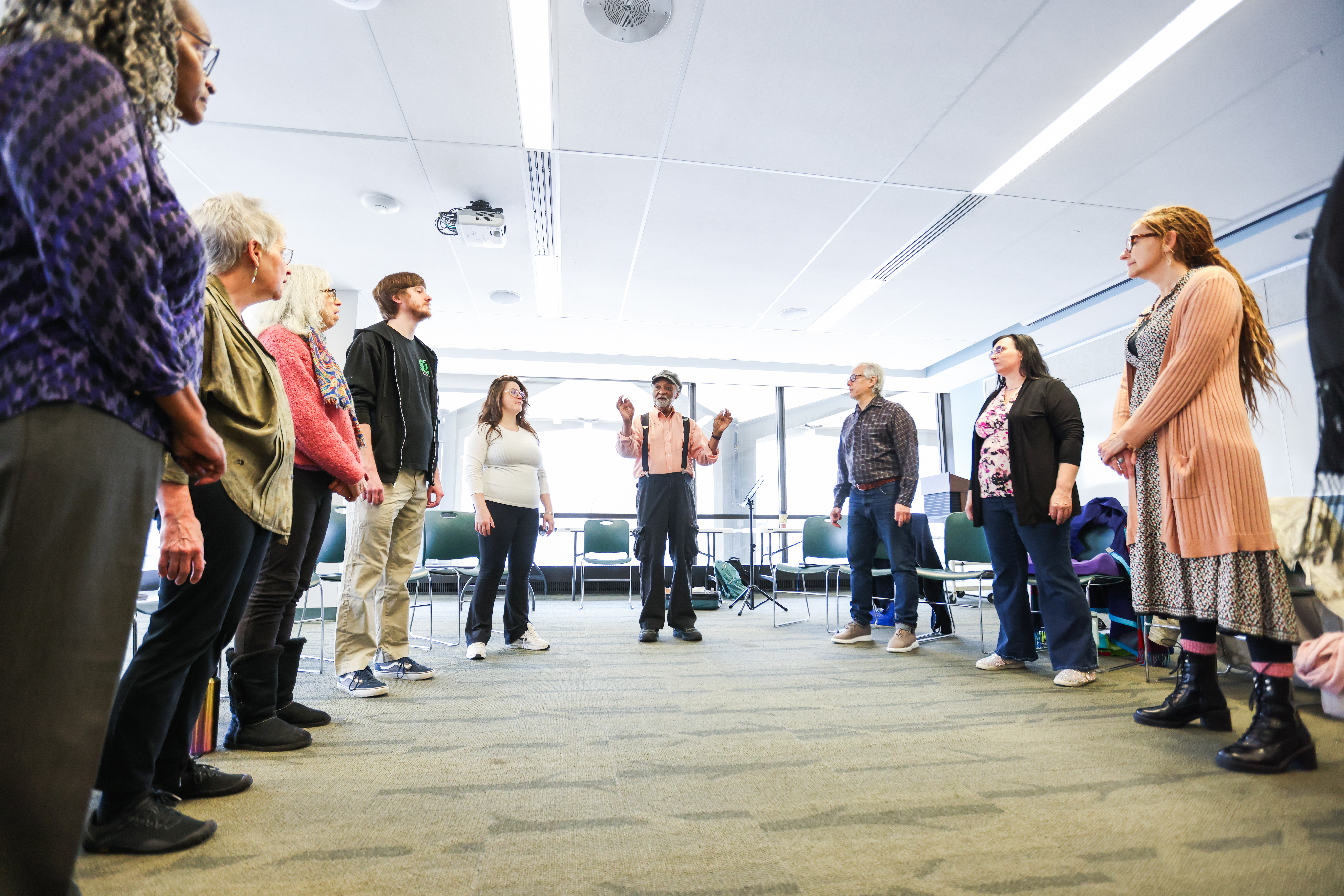 A group of adults stand in a circle in a bright meeting room, listening to a facilitator in the center who gestures as he speaks.