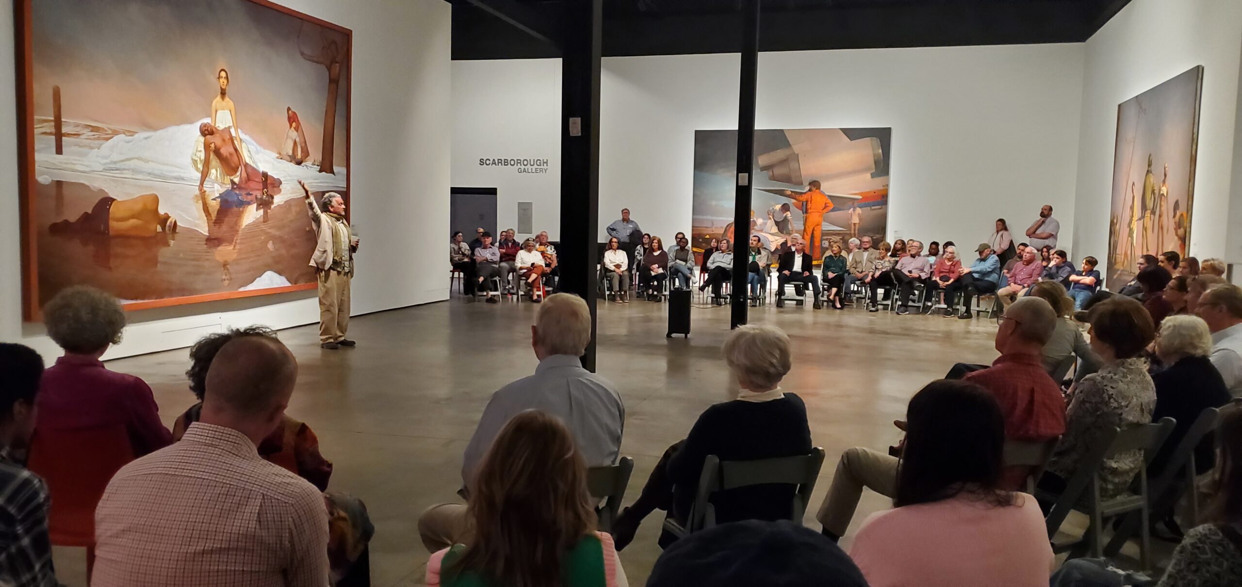 A person presenting to a seated group in an art gallery in front of a large painting.