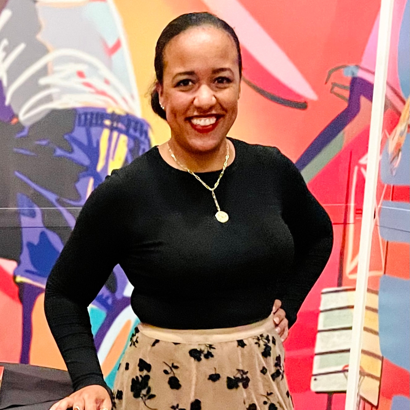 Headshot of a smiling person of medium skin tone wearing a gold necklace, black blouse, and tan skirt.
