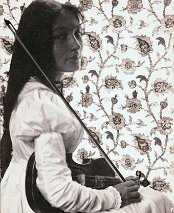 Woman in black and white photo sits to the side holding a violin with floral wallpaper in the background.