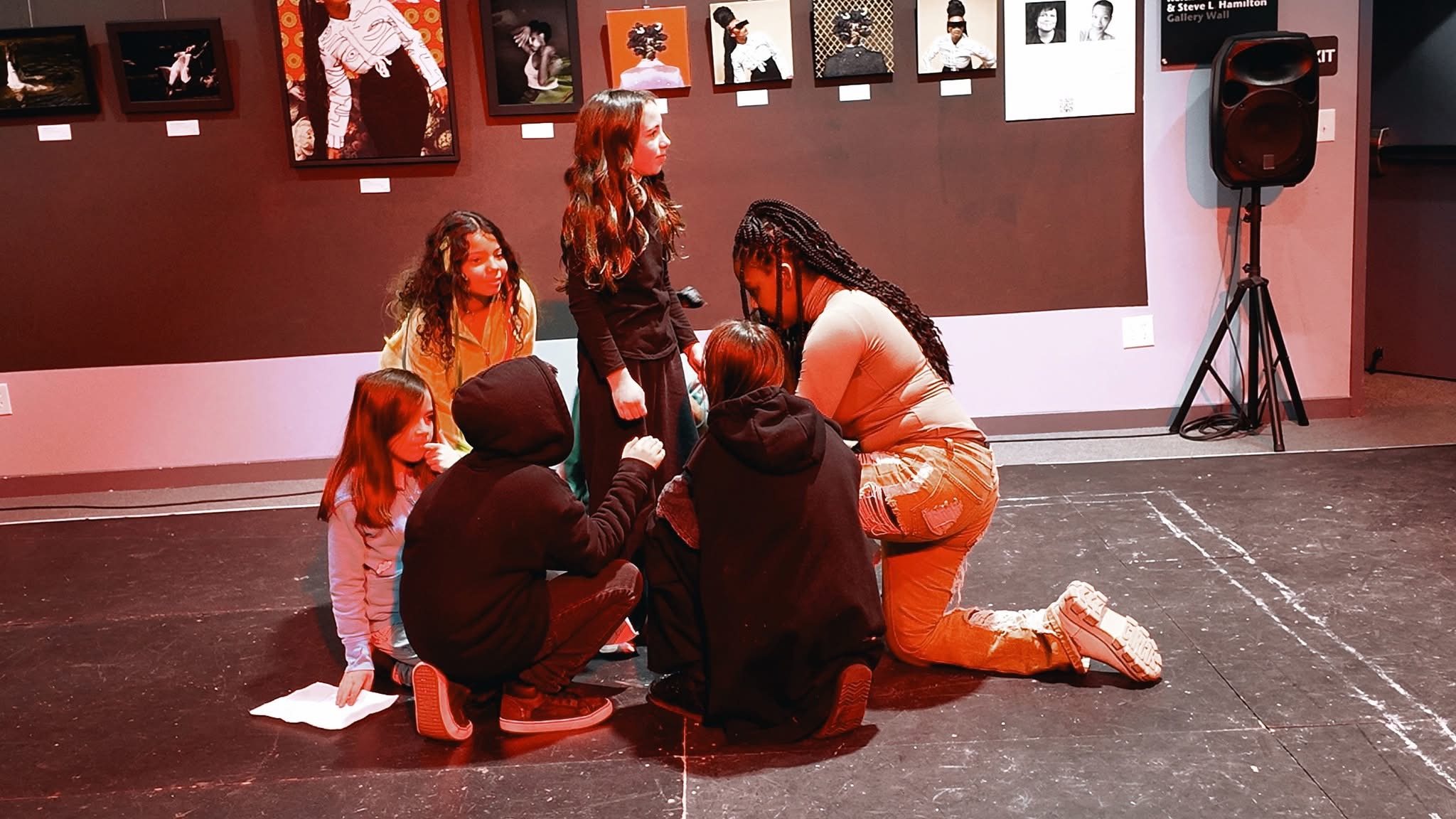 Five kneeling youth in a circle around one standing child in a studio space.