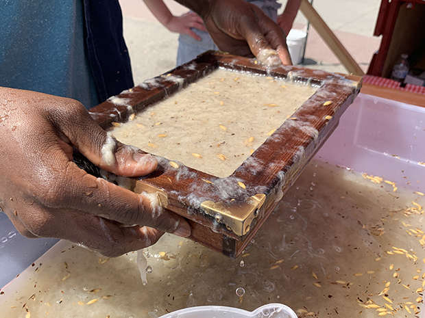 Hands with a dark skin tone hold a wooden frame with wet paper pulp inside.