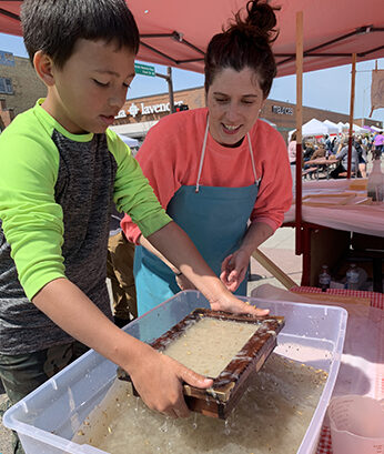 A young person with medium skin wearing a bright green shirt holds a brown frame dunked in paper pulp over a large plastic bin.