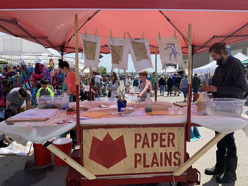 Several people stand under a large pink canopy next to a white folding table with a sign reading 'Paper Plains.'