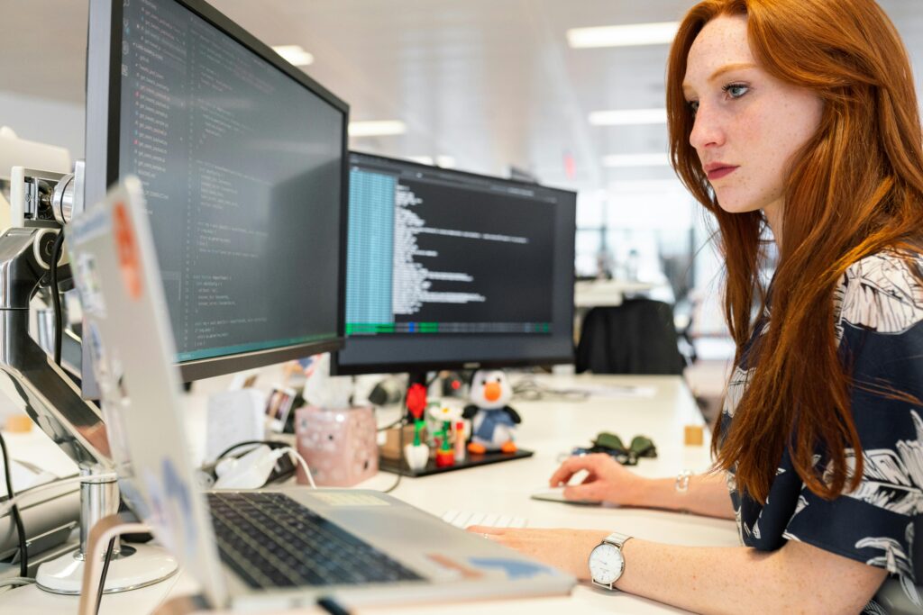 A person with light skin and long ginger hair codes on three monitors