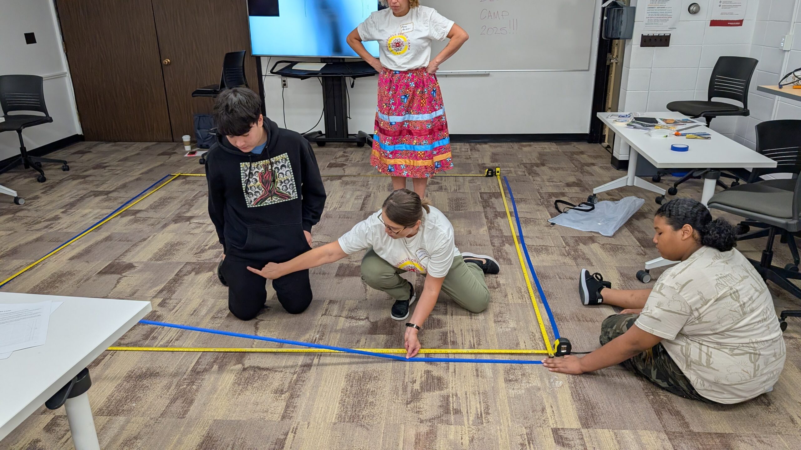 Two young people and an adult work together holding measuring tapes and blue tape to make a large square outline on the floor.