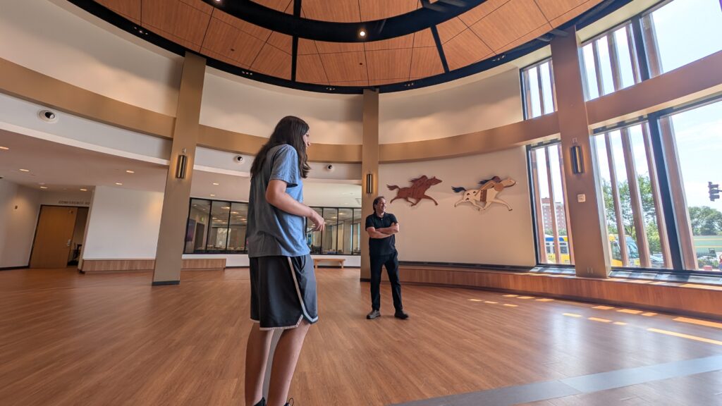 Two people standing in a brightly lit room with large windows and a circular floor plan.