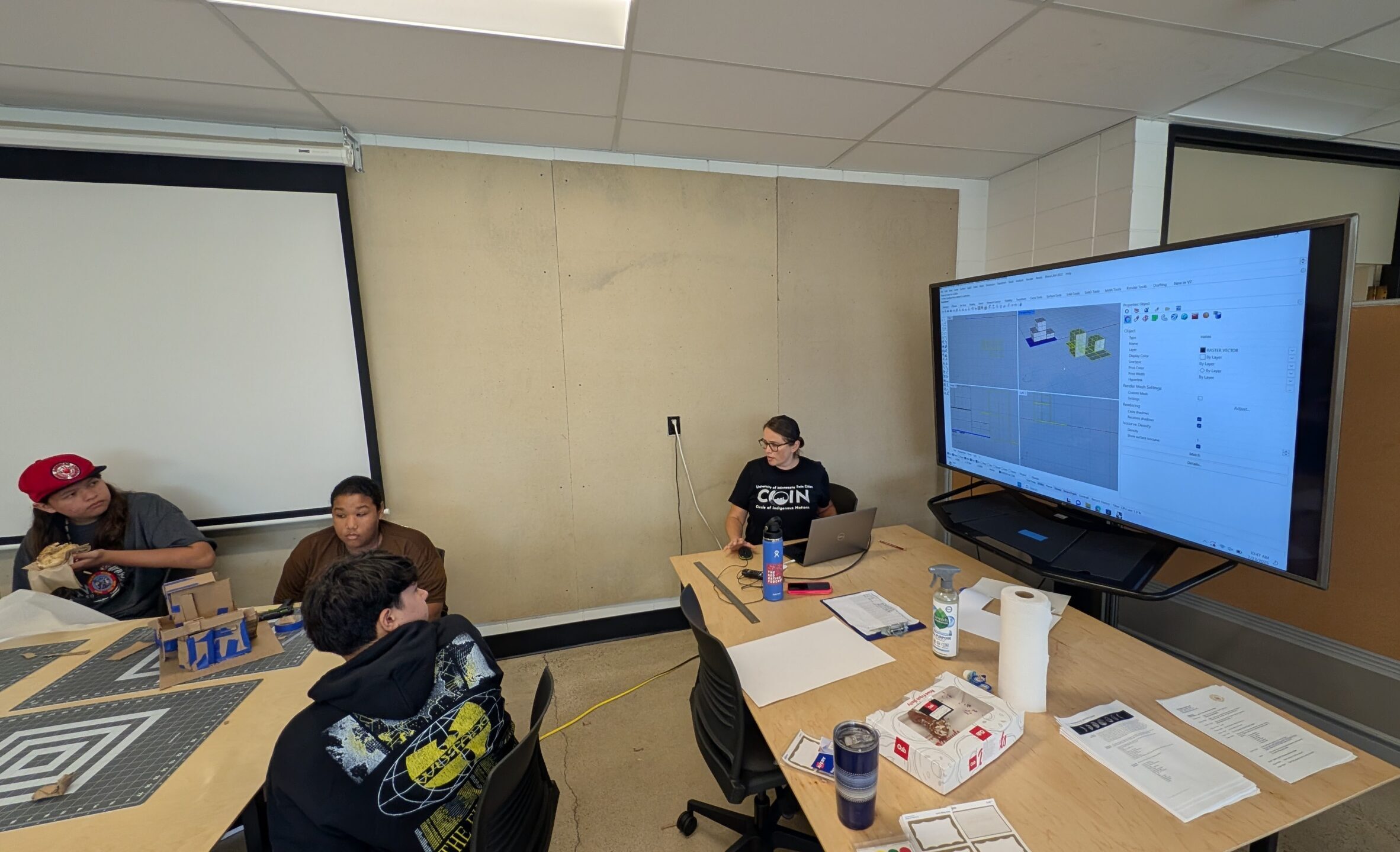 An adult sitting by a big television screen in a classroom as three young people sit at a table nearby.