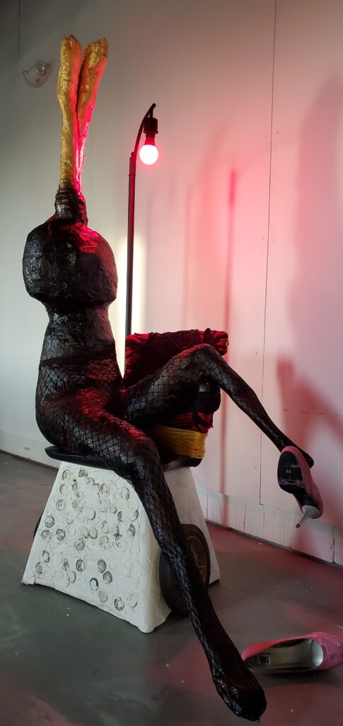 A sculpture of a woman sitting on a chair illuminated by a red light. Where her head should be is a giant wick-like object. High heels fall off of her feet.