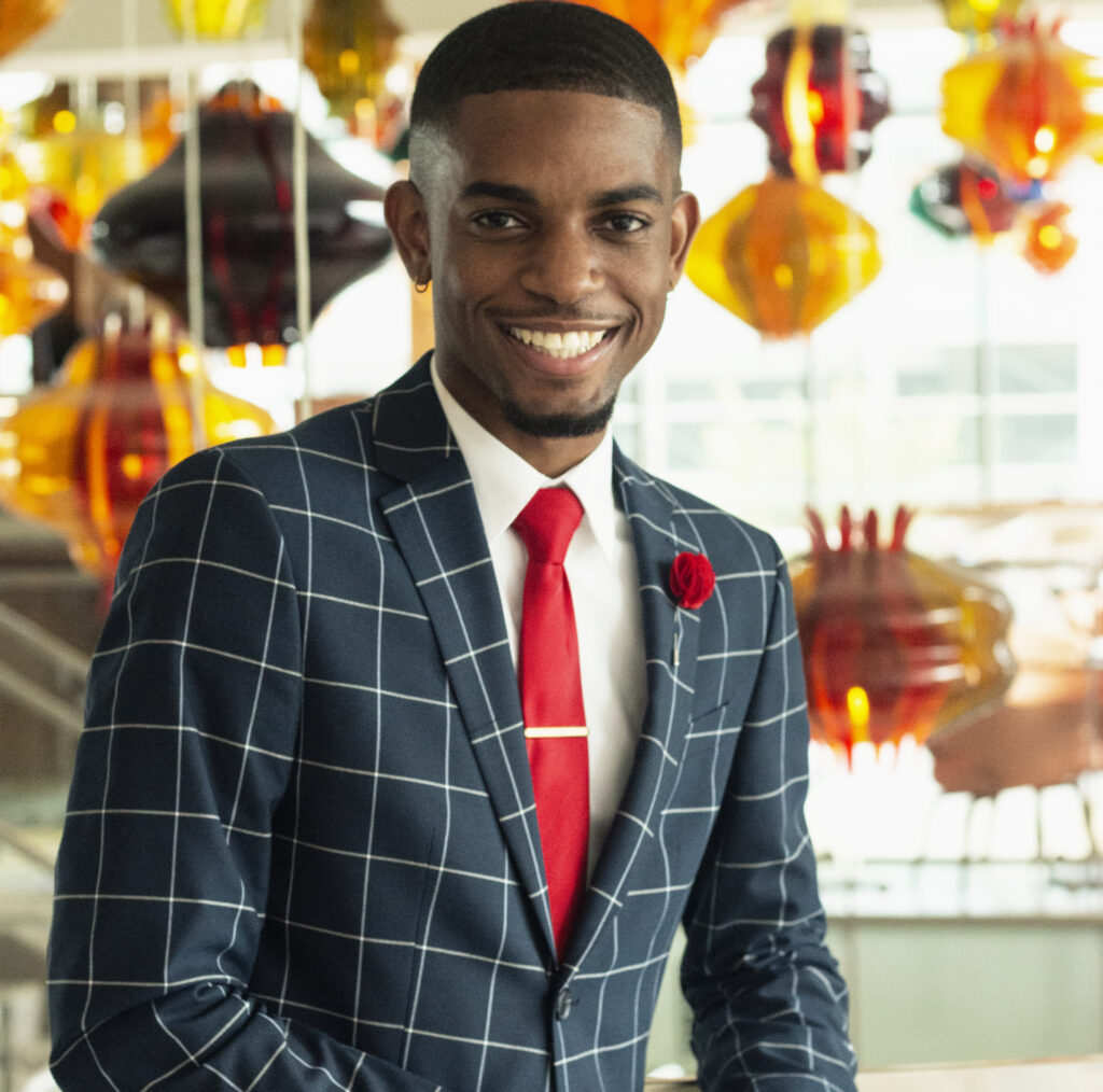 Headshot of a smiling person of dark skin tone and very short black hair and beard/mustache, wearing a striped suit jacket with a red tie and flower. In the background many gold and red glass light fixtures hang from the ceiling.