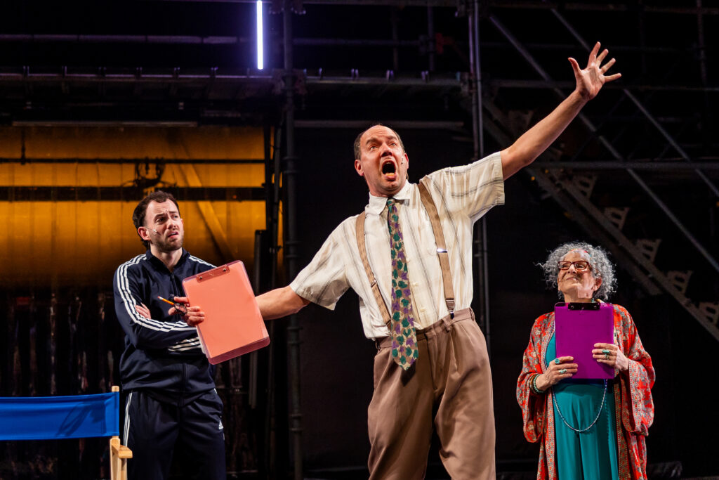Three people on stage, one holding a clipboard and exclaiming with their whole body.