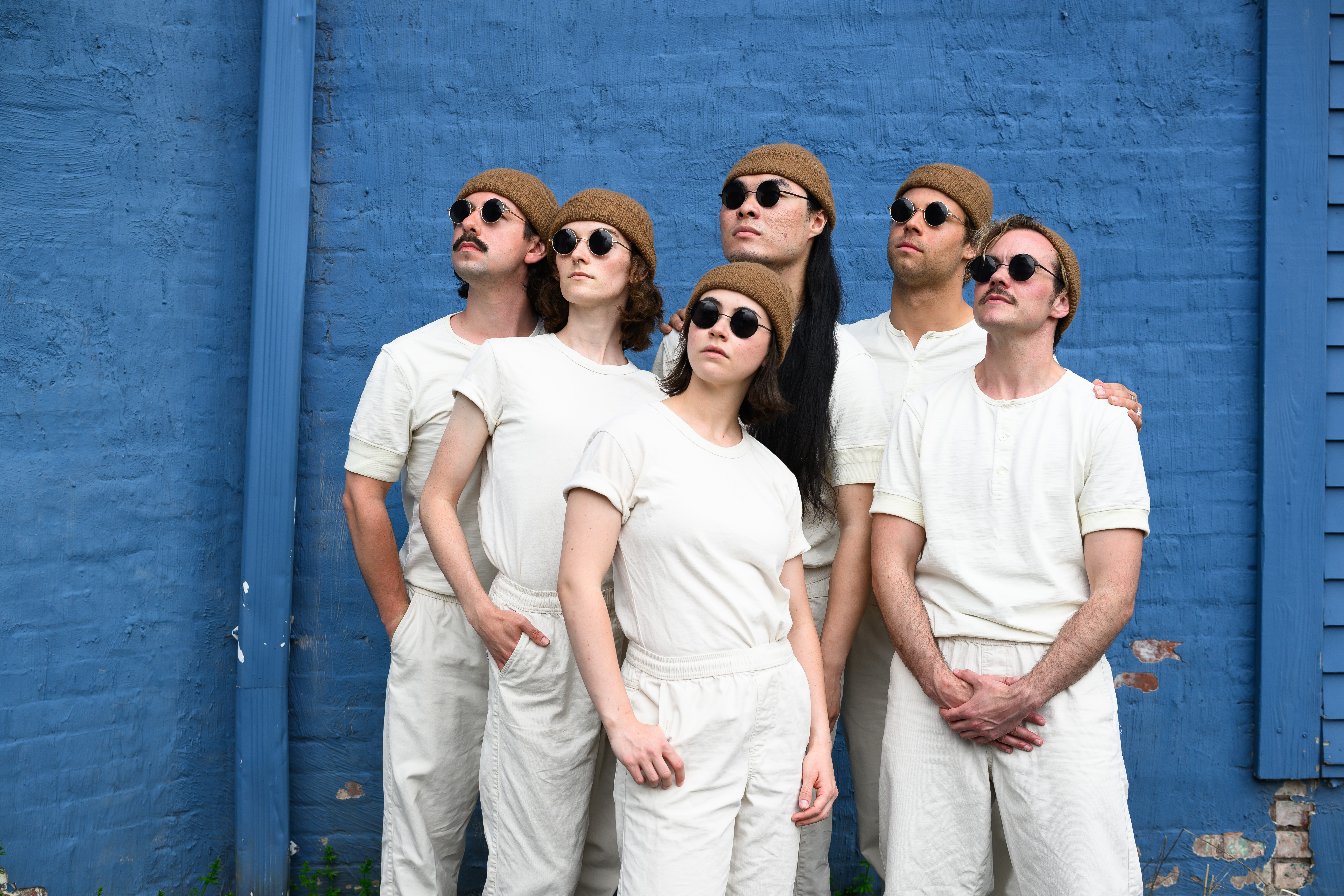 A group of six people in white costumes, beanies, and sunglasses.