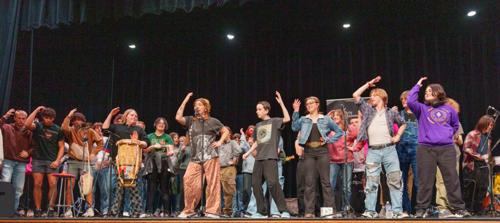 A diverse group of students and musicians stand on stage, mimicking dance movements during an energetic school assembly performance.