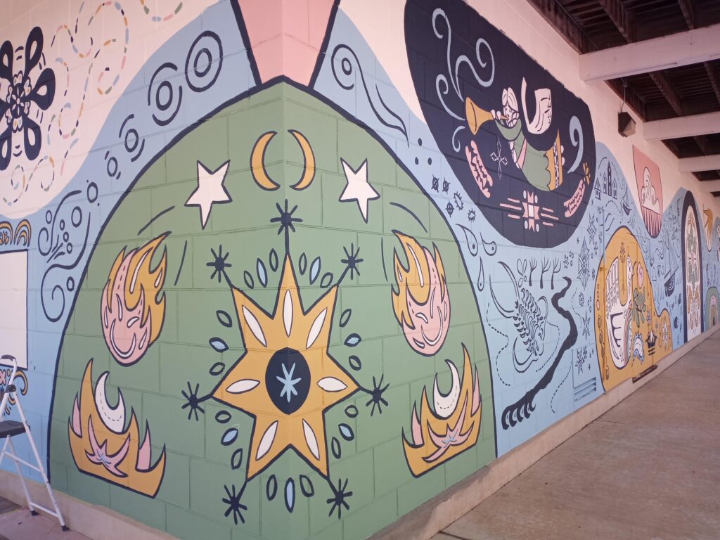 A mural painted on the side of a building. Pastel colors and bold lines show stars, meteors, angels, sea life, and other abstract forms.