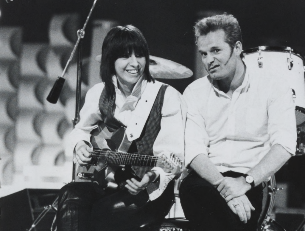 A woman and a man sit on a stage; the woman smiles and plays guitar; the man makes a silly face and looks at the camera in a black and white image.