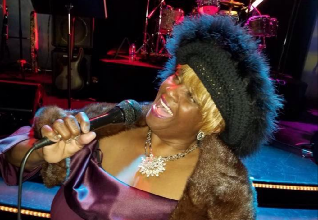 Dark-skinned woman with blonde hair and a fuzzy black hat sings on stage.