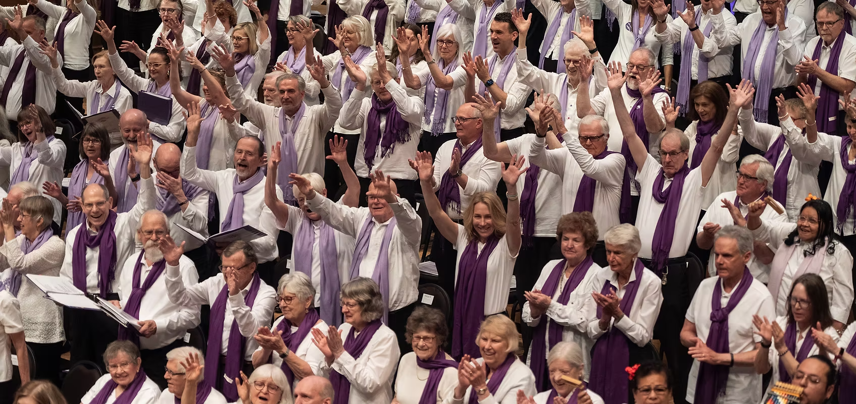 A crowd of people wearing white shirts and purple scarves clap, raise their hands, and hold books.