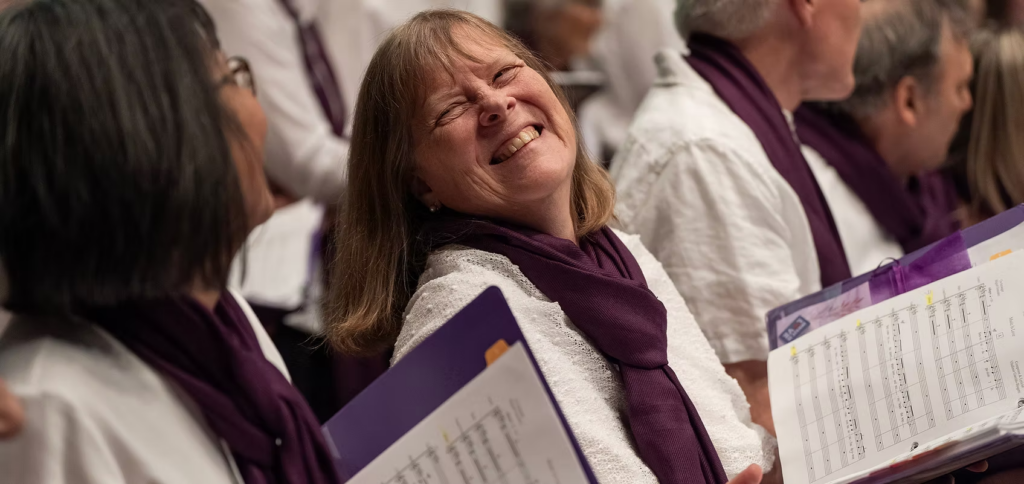 A light-skinned person wearing a white shirt and purple scarf smiles and looks at another person while holding a binder with sheet music inside.