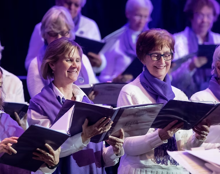 People smile and hold black binders while wearing white shirts and purple scarves against a dark background.