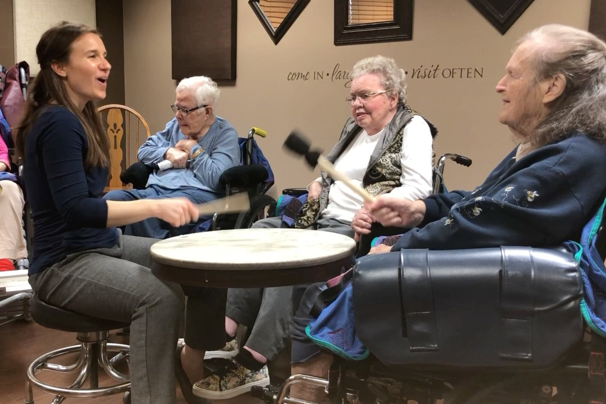 Three light-skinned people sit around a drum with mallets.