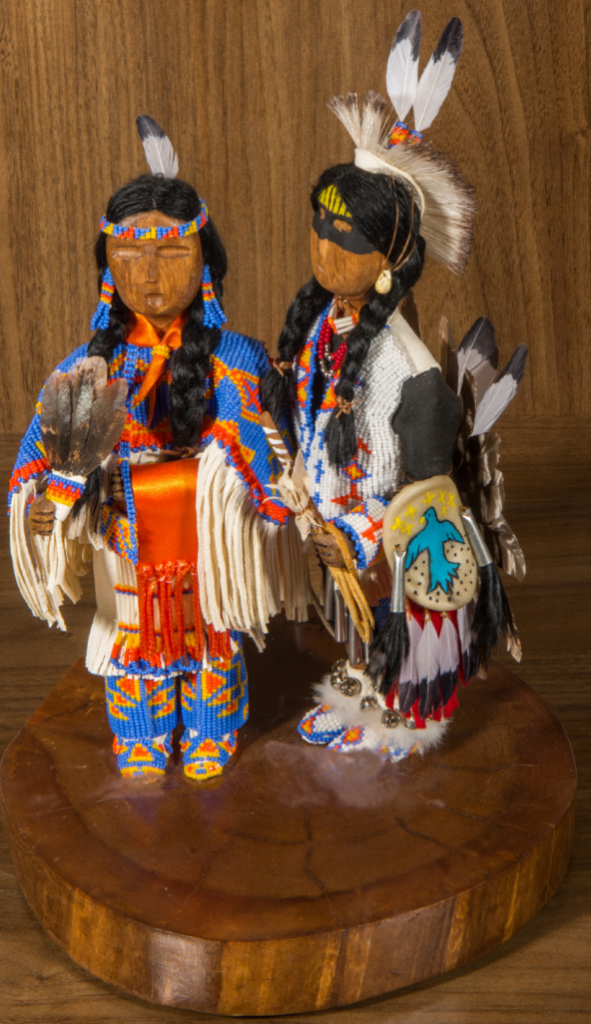 Two figures made of various materials standing on a slice of wood.