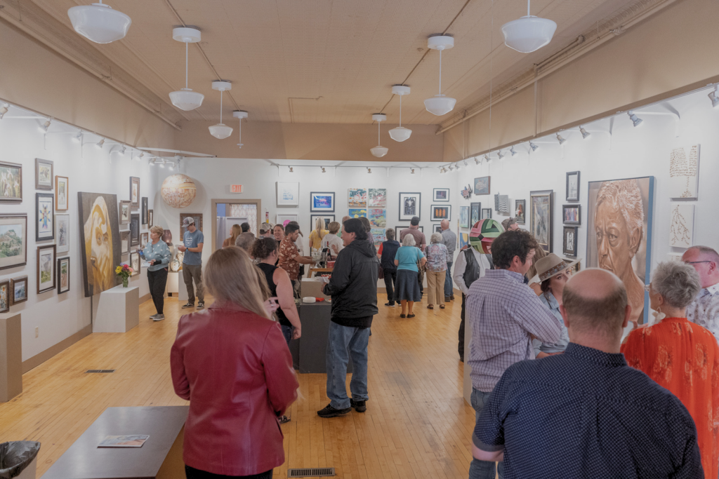 Many people gather in a well lit art gallery, observing artwork on the walls and talking to one another.