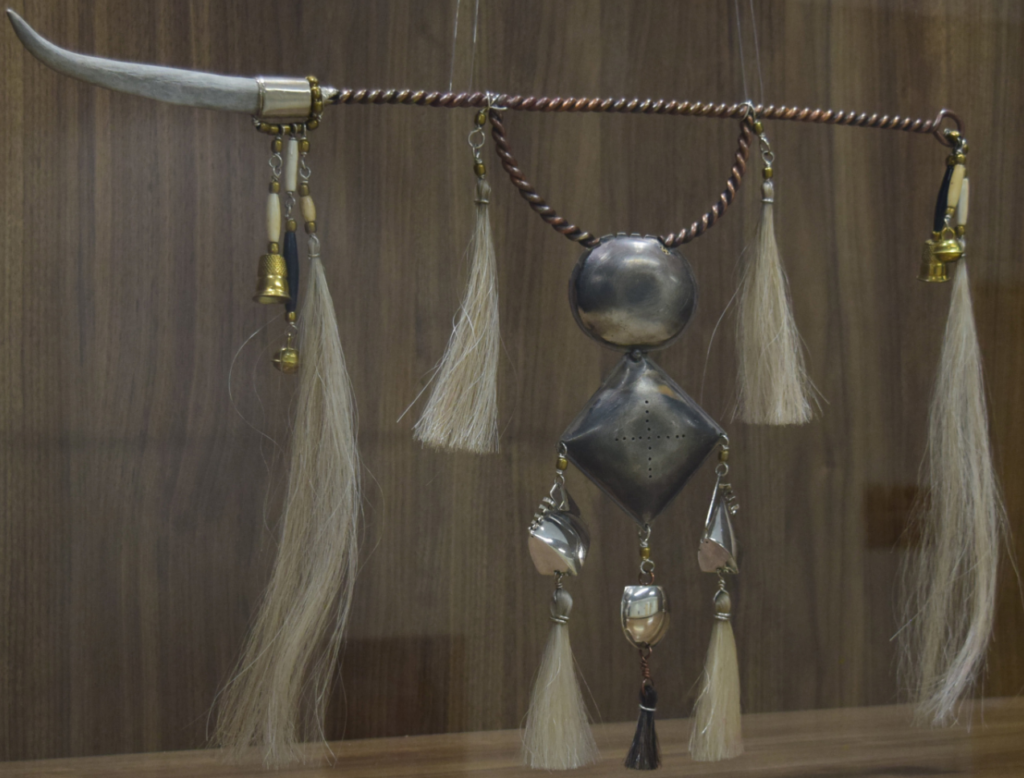 A gray sculpture with various materials, tassels, and spheres.