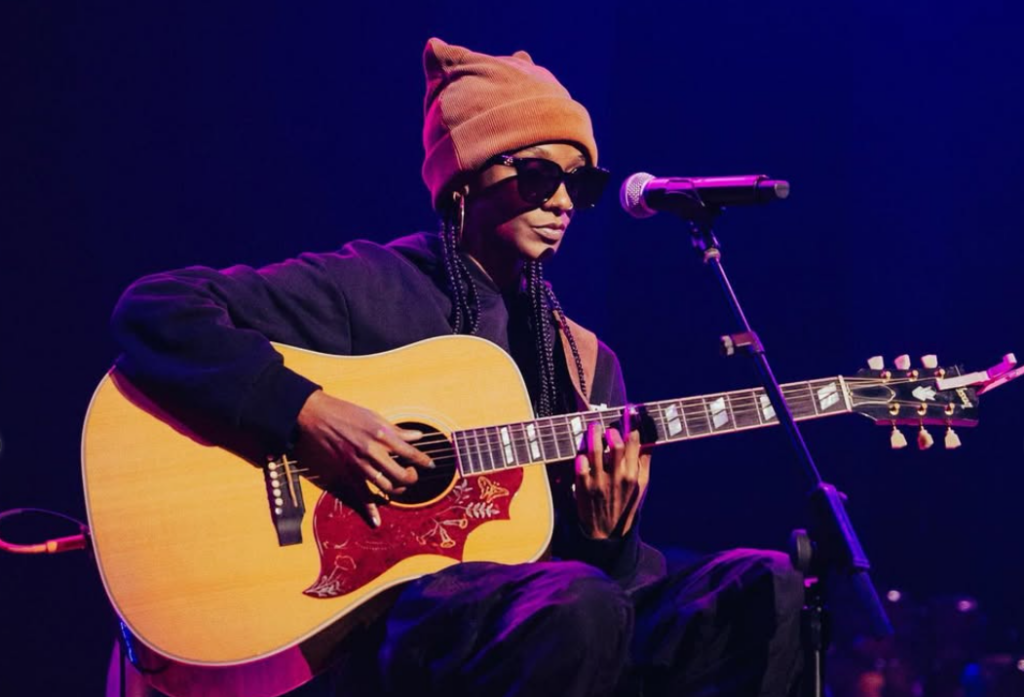 Dark-skinned woman wears a beanie and sunglasses on stage and plays guitar.
